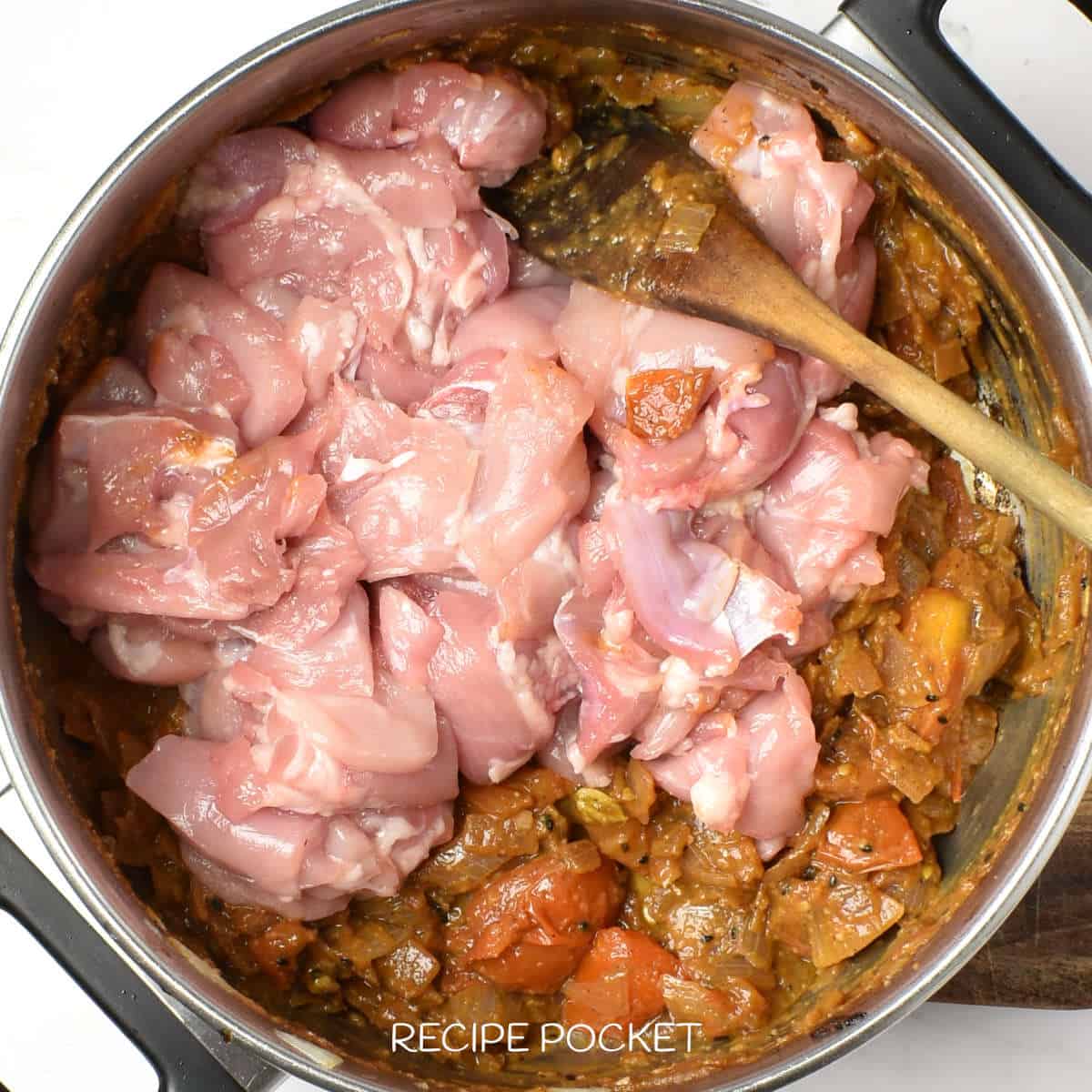 Raw chicken in a pot with cooked onions and tomatoes.