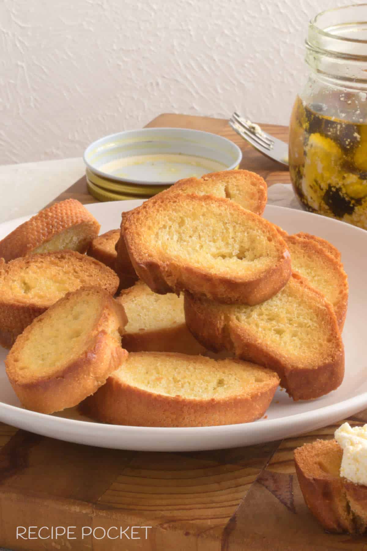 A plate of freshly made baguette crostini with a jar of feta in oil in the background.