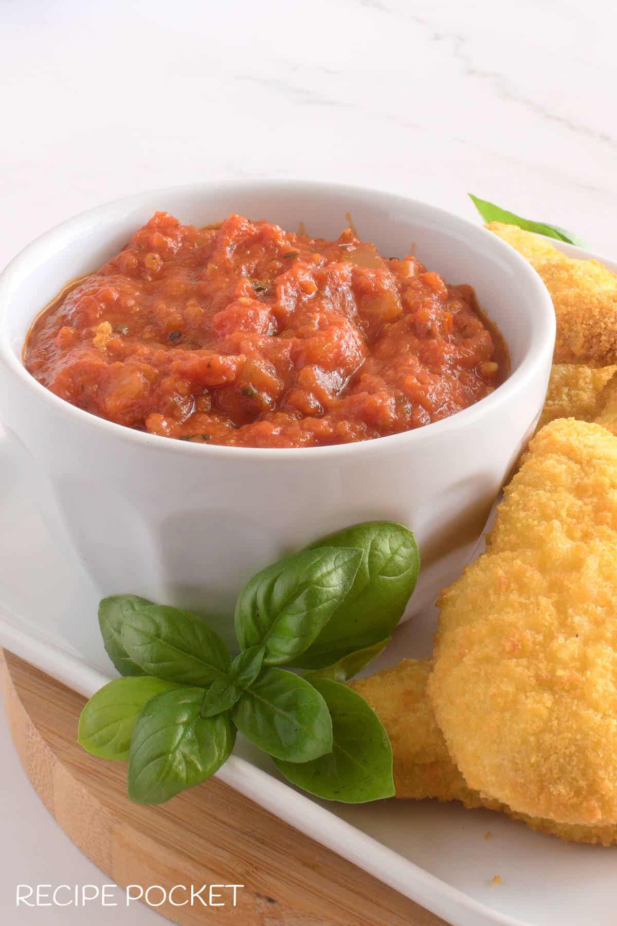 A white bowl with marinara dip and crumbed chicken pieces on the side.