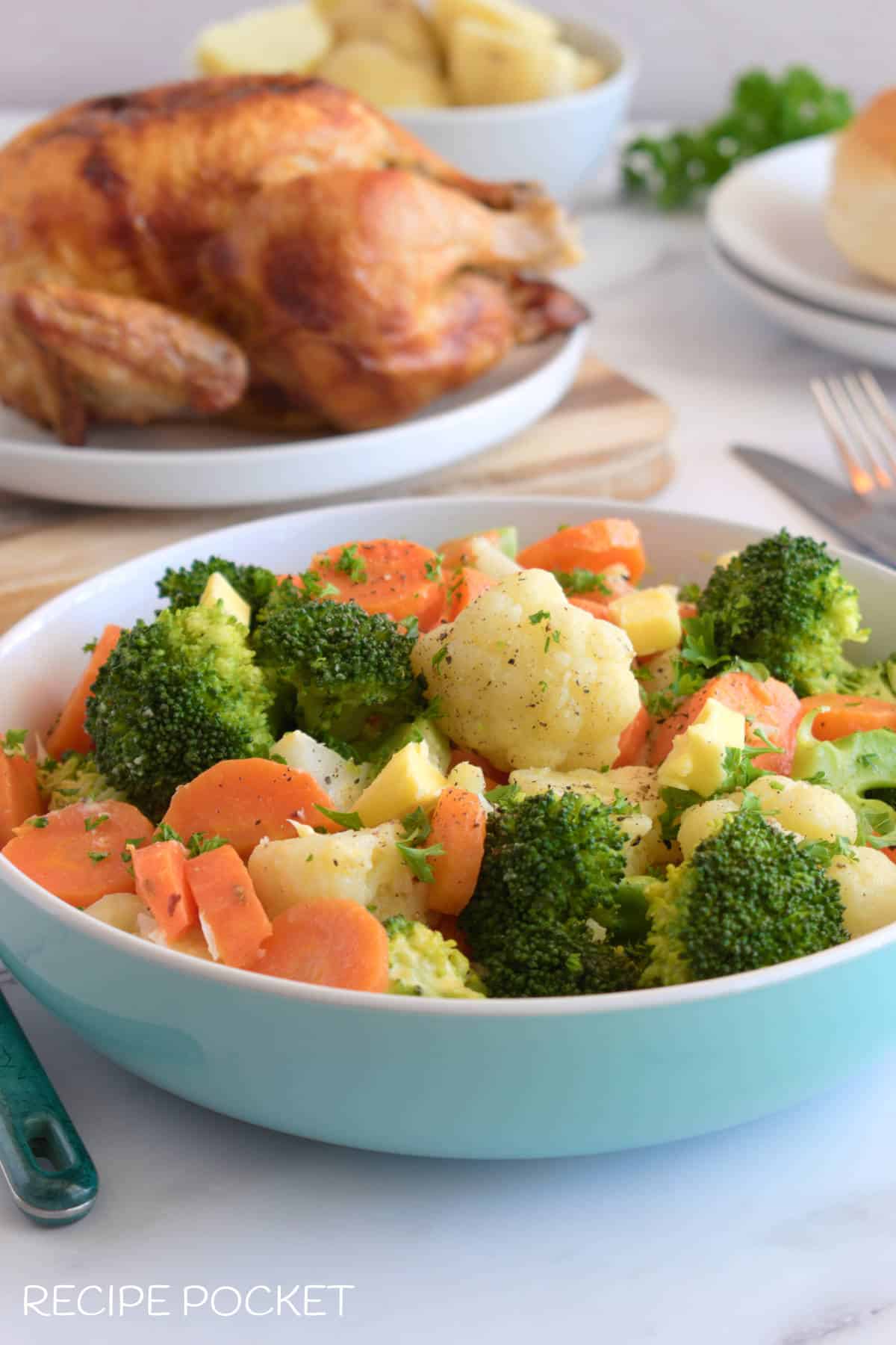 Buttered vegetables in a blue serving dish with roast chicken in the background.
